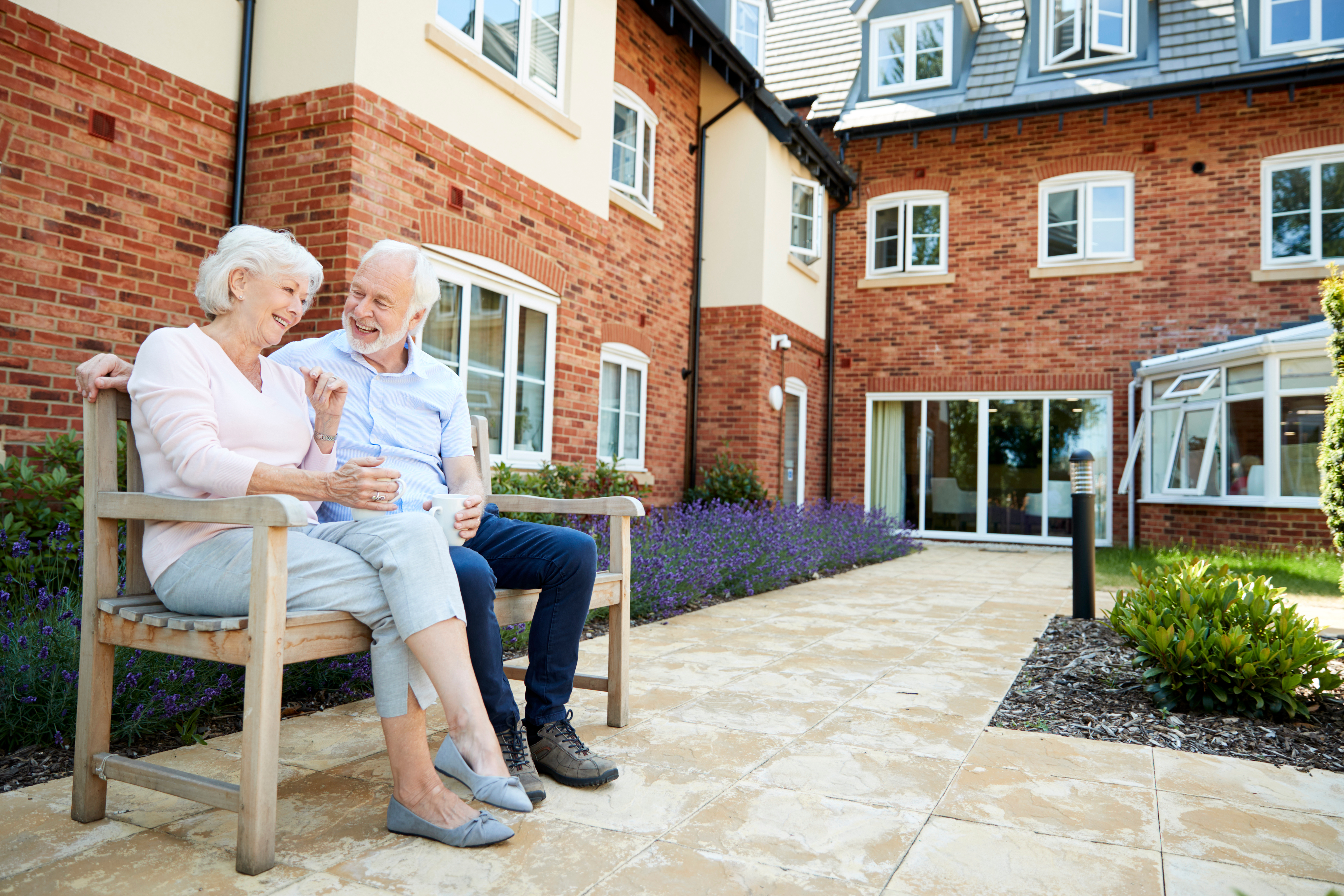 Retired Couple Sitting On Bench With Hot Drink In Assisted Living Facility Ein glückliches älteres Paar auf einer Parkbank