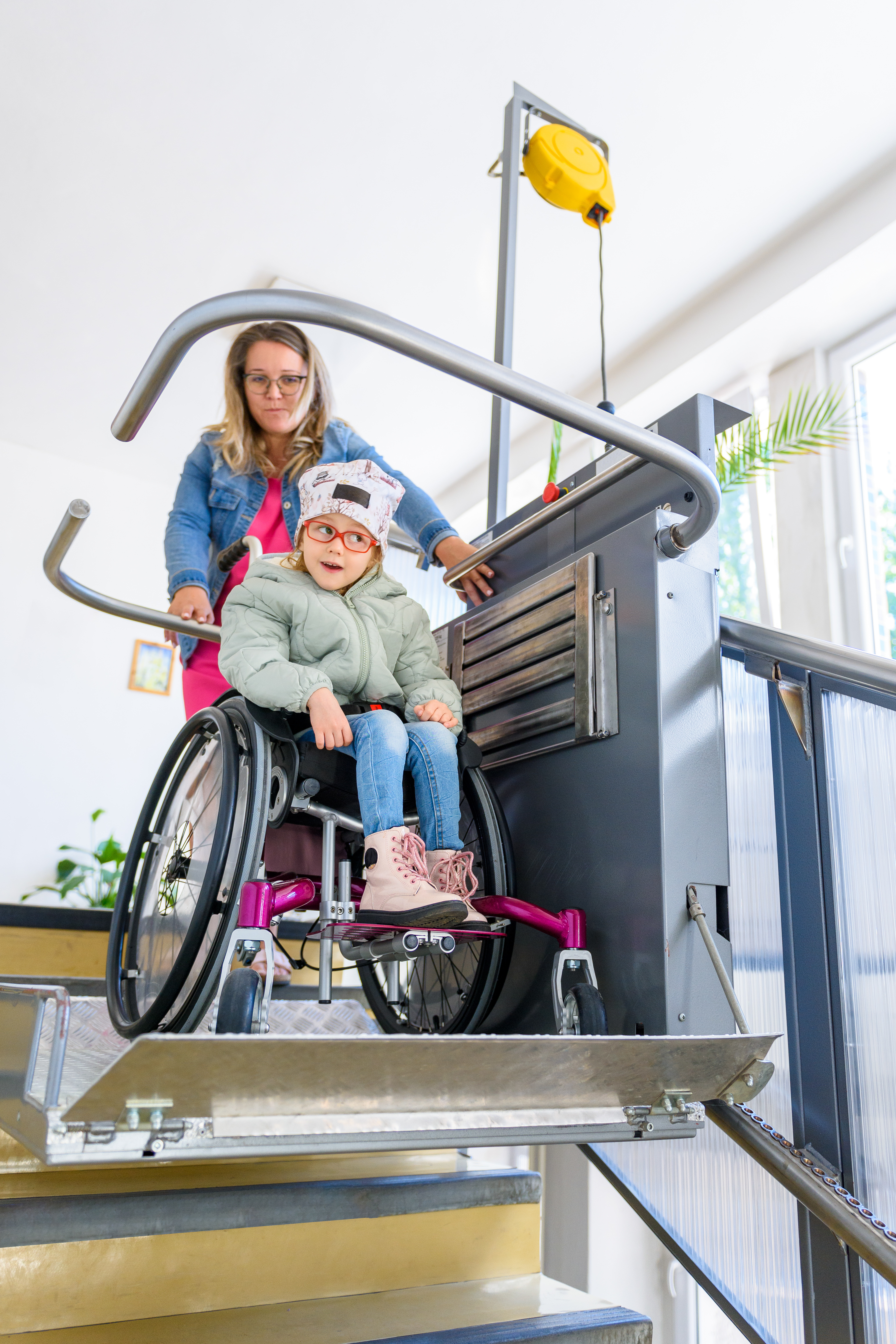 Mother with a young child living with cerebral palsy using electric wheelchair lift to access public building. Special lifting platform for wheelchair users. Disability stairs lift facility.